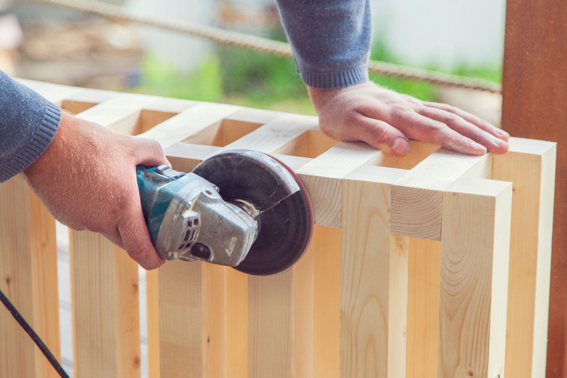Crop anonymous woodworker using electric angle grinding machine to polish wooden construction on sunny backyard